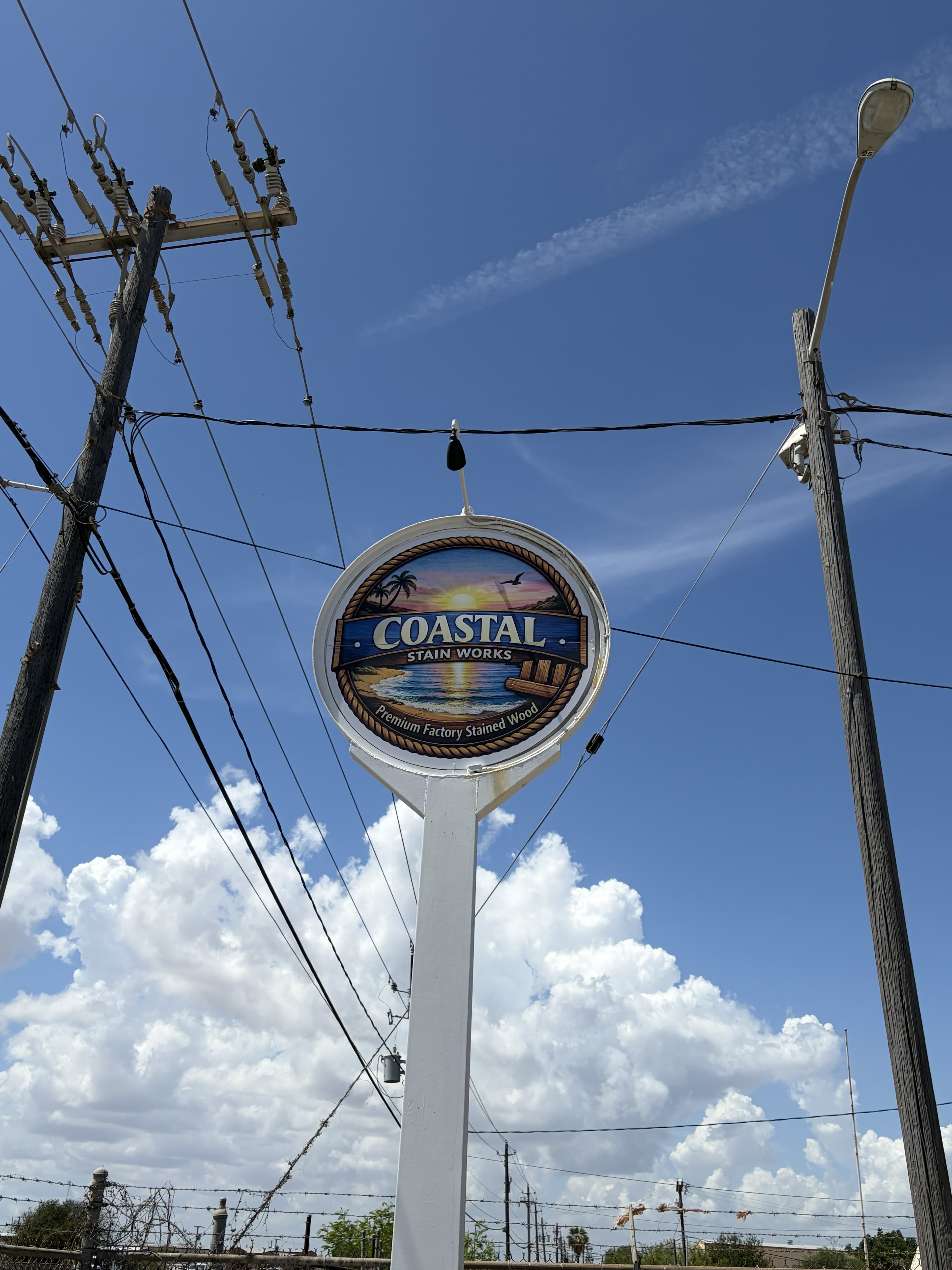 Circular Coastal Stain Works sign featuring a beach sunset, set against a bright cloudy sky.